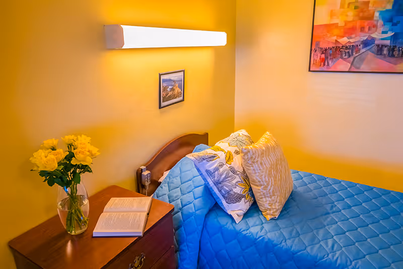A cozy bedroom with a single bed dressed in a blue quilt, decorative pillows, a bedside table with a vase of yellow roses and an open book, and wall art under a wall light.