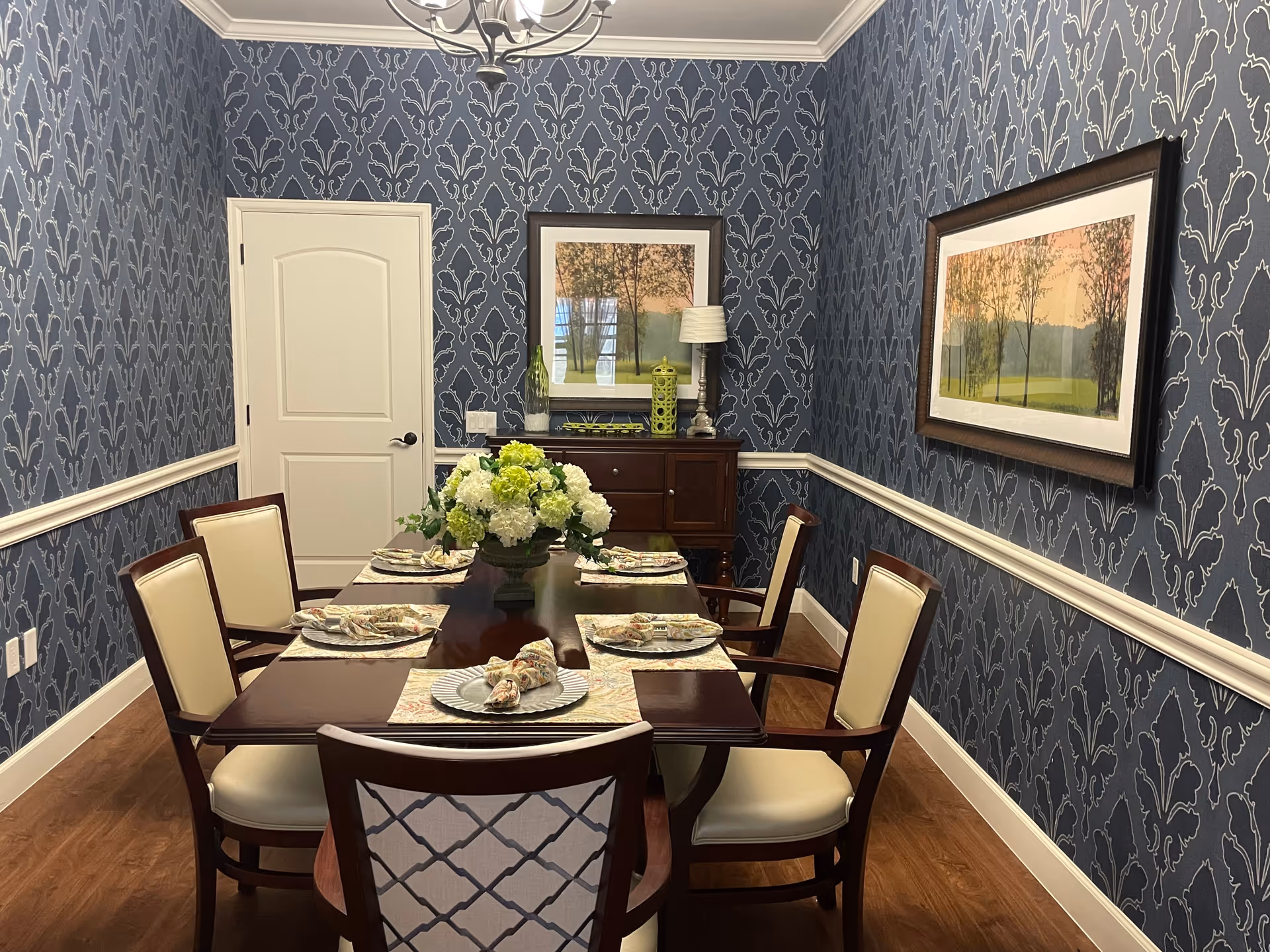 A formal dining room with a dark wooden table set for six people. The table has placemats, plates, and napkins arranged neatly, with a centerpiece of white and green flowers. The room features blue patterned wallpaper with white trim, two framed landscape paintings on the walls, a wooden sideboard with decorative items, and a chandelier hanging from the ceiling.