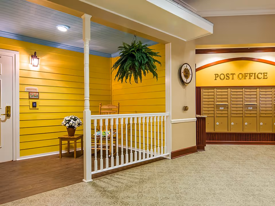 Indoor common area with yellow paneled wall, white railing, rocking chair, hanging fern and a bank of mailboxes labeled "POST OFFICE".