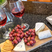 A wooden board with clusters of red grapes, three types of cheese, and round crackers on a dark granite countertop. Two glasses of red wine are placed behind the board.
