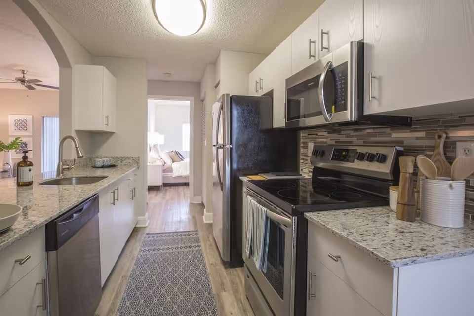 Modern kitchen with granite countertops, stainless steel appliances including a refrigerator, microwave, and electric stove. There is a sink with a faucet on the left side and kitchen utensils in a white container on the right countertop. A patterned rug runs along the wooden floor. In the background, a bedroom with a bed and lamp is visible through an open doorway.