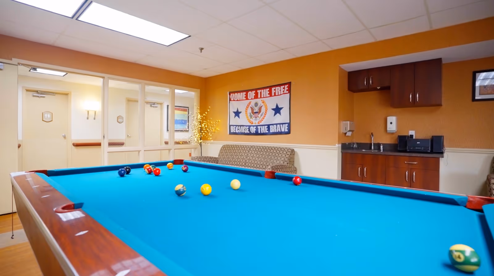 A bright interior recreation room with a blue-felt pool table in the foreground, a couch and small kitchenette against the far wall, and a patriotic 'Home of the Free' sign.