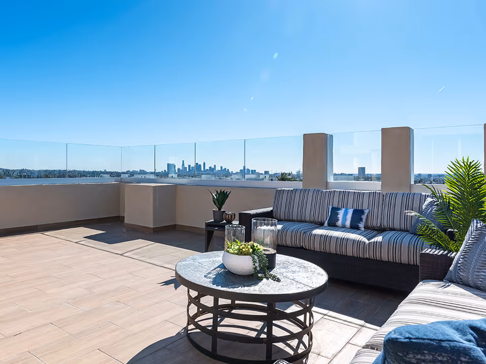 Outdoor rooftop patio area with modern wicker sofas featuring striped cushions and blue accent pillows, a round metal coffee table with decorative plants, glass railing offering a clear view of a city skyline under a bright blue sky.