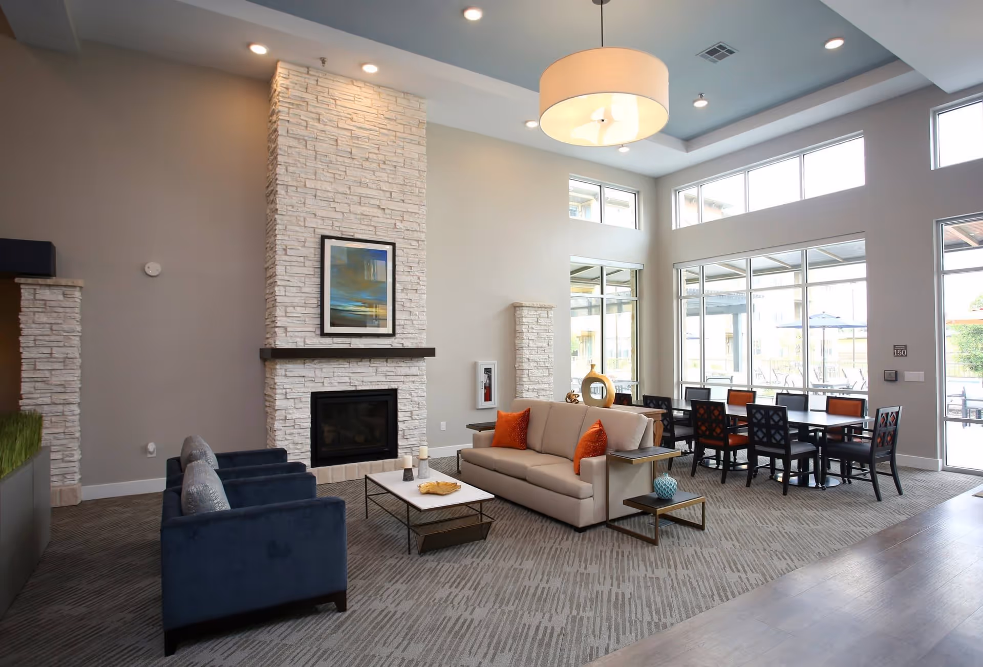 A spacious and modern living room area with a beige sofa adorned with orange pillows, two dark blue armchairs, a white coffee table, and a stone fireplace with a framed artwork above it. Large windows let in natural light, and a dining table with chairs is visible in the background near the windows.