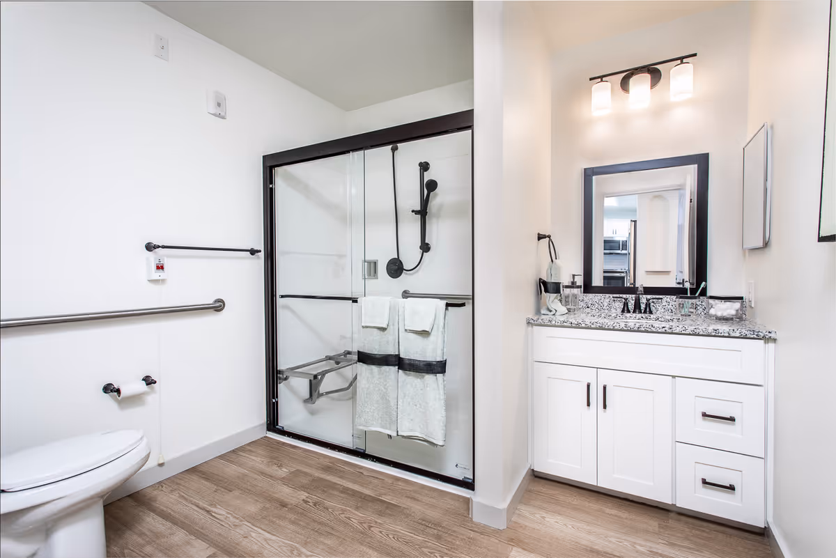 A modern bathroom with a glass-enclosed shower featuring a black frame and a mounted showerhead. There are two white towels hanging on the shower door. The bathroom also includes a white toilet with a grab bar next to it, and a white vanity with a granite countertop, a sink, and a mirror above it. The floor is wood-style, and the walls are painted white.
