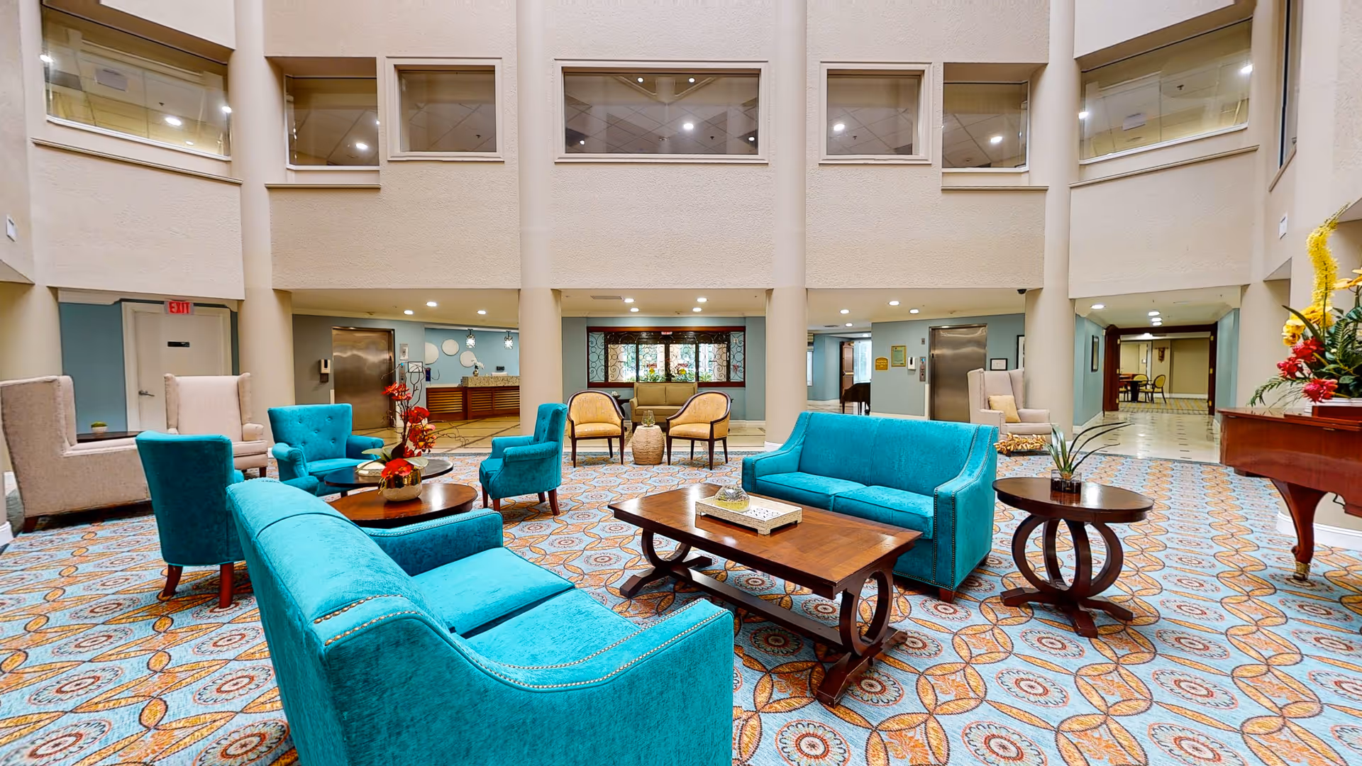 A spacious and well-lit common area with turquoise sofas and armchairs arranged around wooden coffee tables on a patterned carpet. The room has high ceilings with multiple windows on the upper walls, beige pillars, and light-colored walls. There are additional beige armchairs and a piano with a floral arrangement on the right side.