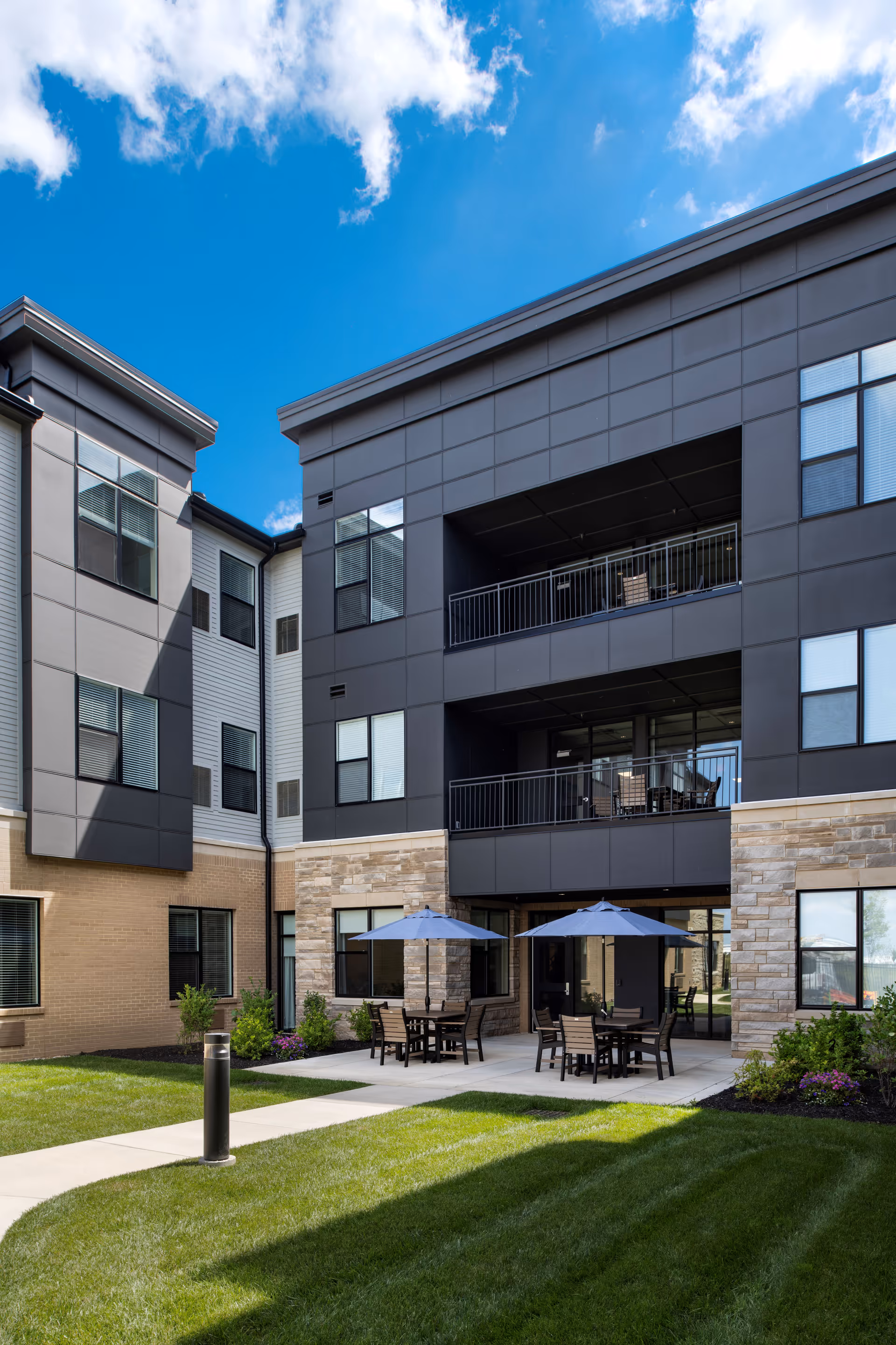 Outdoor patio area at Charter Senior Living of Gateway Park with tables, chairs, and blue umbrellas on a concrete surface surrounded by green grass and landscaping, adjacent to a modern multi-story building under a blue sky with some clouds.