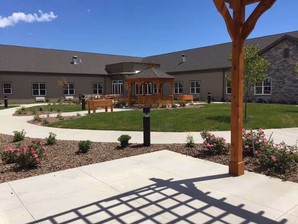 Sunlit courtyard with a wooden gazebo, paved walkways, flower beds, and the surrounding single-story building.