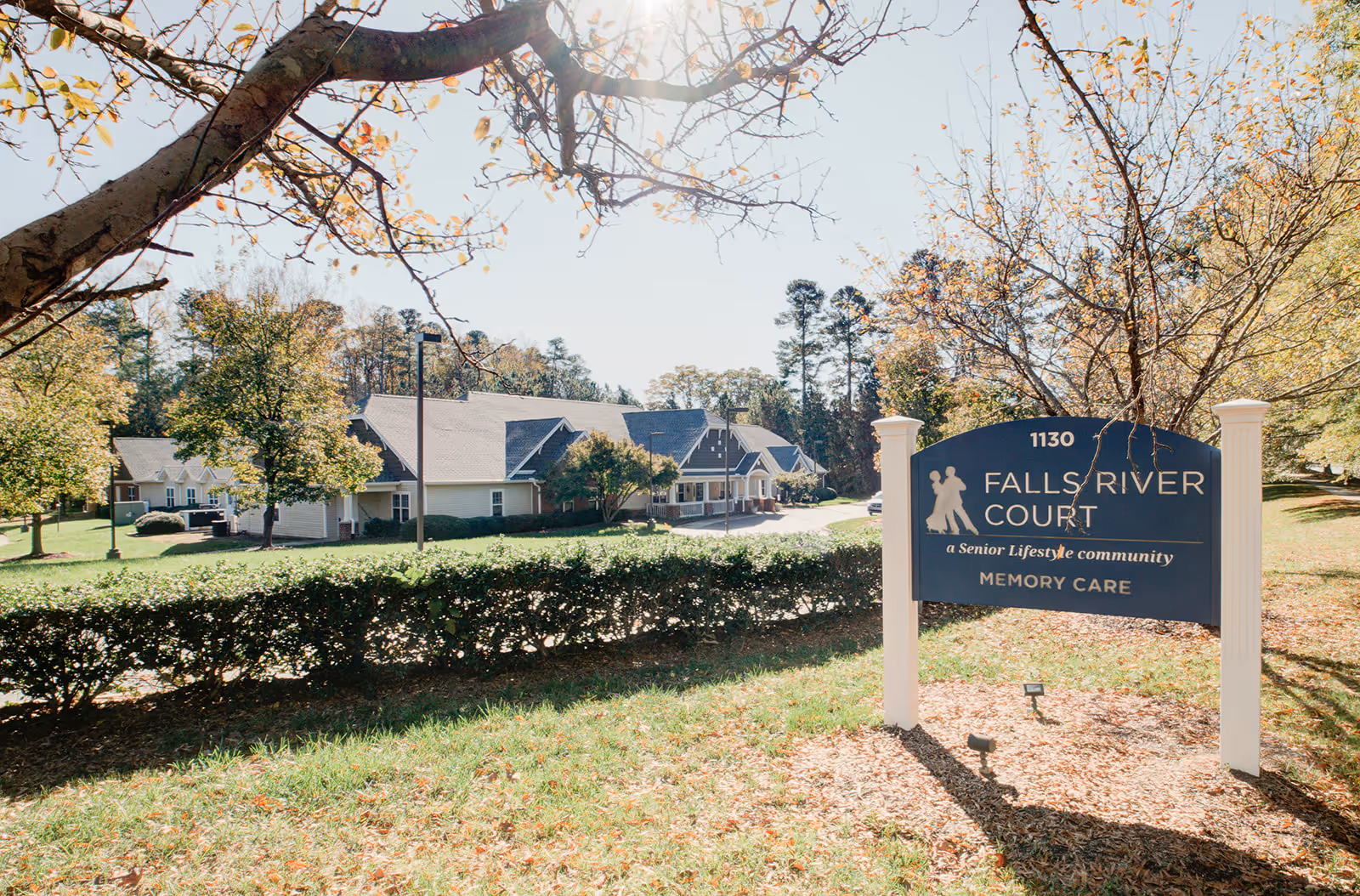 Outdoor view of Falls River Court Memory Care facility with a large sign in the foreground displaying the facility name and description. The building is surrounded by trees and greenery under a clear sky.
