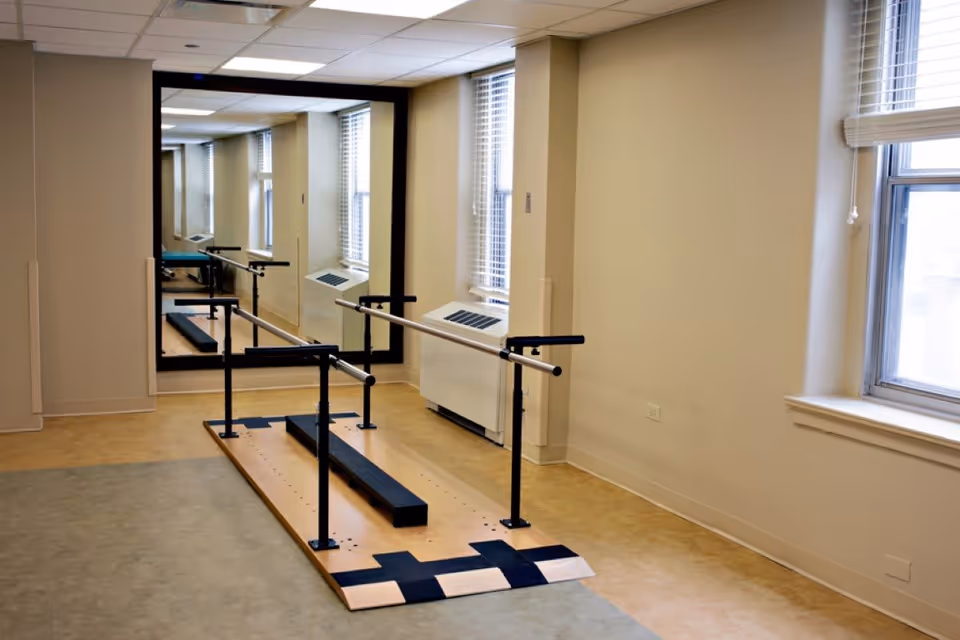 A rehabilitation or physical therapy room with parallel bars mounted on a wooden platform for walking exercises. The room has beige walls, a large mirror on one wall, and windows with blinds letting in natural light.