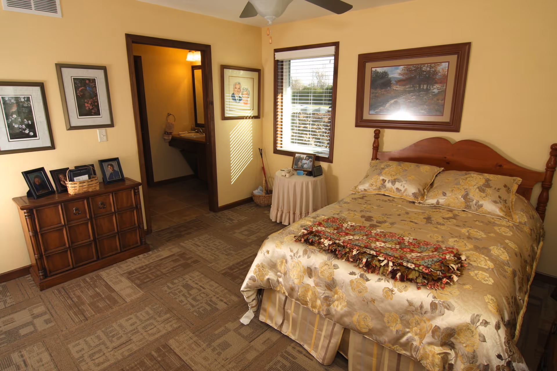 A cozy bedroom with a wooden bed frame and a floral patterned bedspread in shades of yellow and beige. A colorful folded blanket lies at the foot of the bed. To the right of the bed is a small round table covered with a white cloth, holding a framed photo, a clock, and a telephone. A window with white blinds lets in natural light. On the left wall, there is a wooden dresser with framed photos and a basket on top. The walls are painted a warm beige color and decorated with framed pictures. An open doorway leads to a bathroom with a sink and mirror visible. The floor is carpeted with a patterned design, and a ceiling fan with a light fixture hangs above.