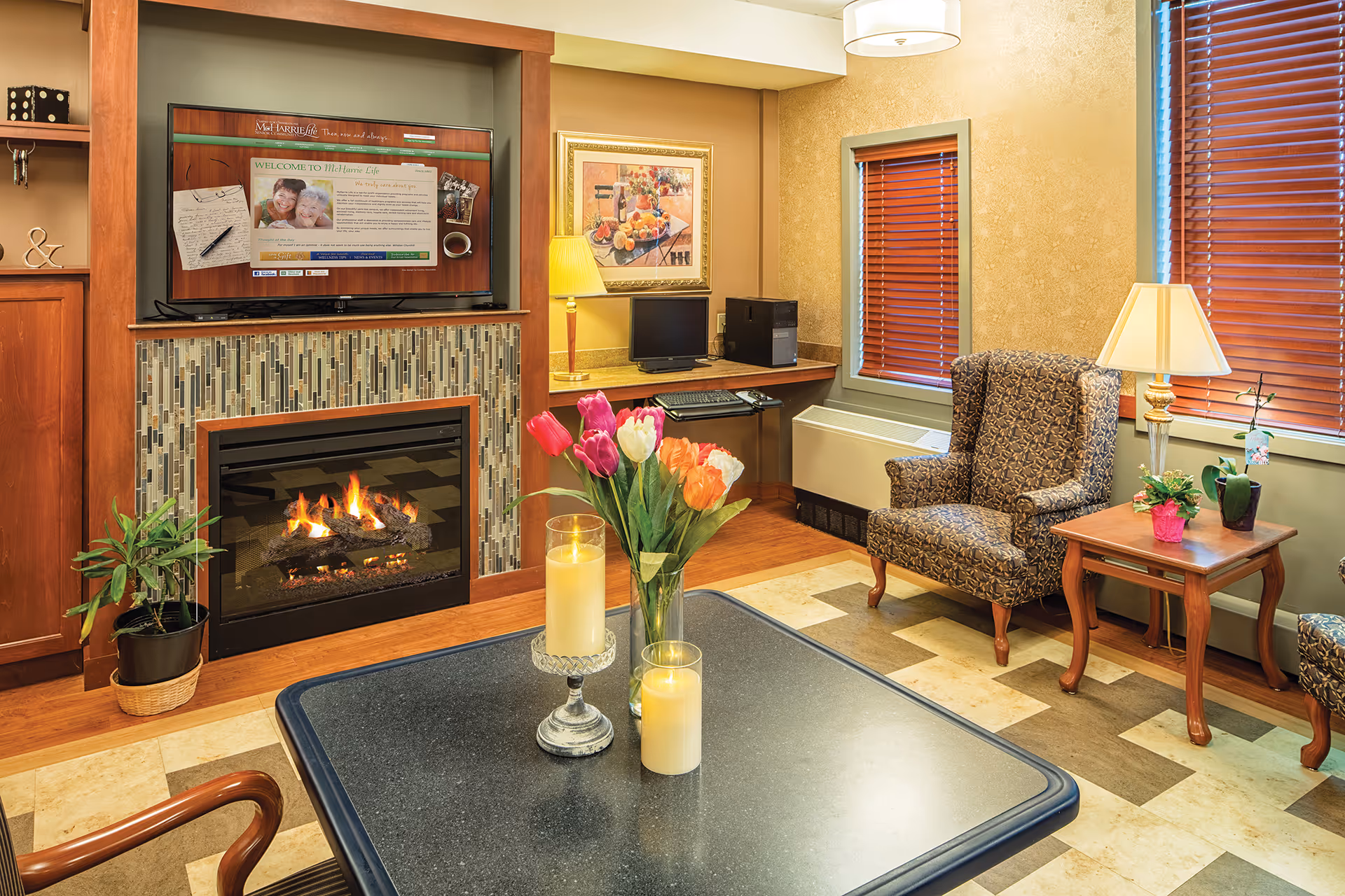 Cozy living room area with a lit fireplace surrounded by mosaic tiles, a flat-screen TV mounted above it displaying a welcome message from Mc Harrie Pointe, a patterned armchair, a wooden side table with a lamp and plants, a desk with a computer, and a table in the foreground with candles and a vase of colorful tulips.