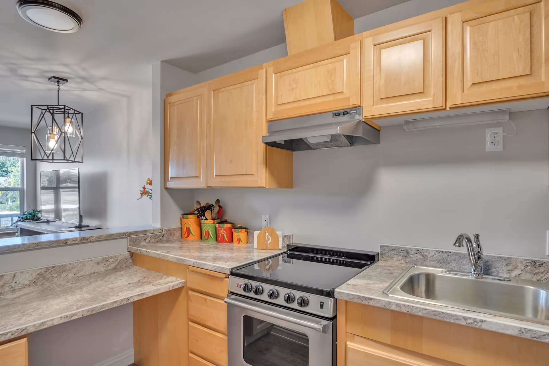 A modern kitchen with light wood cabinets, a stainless steel stove and oven, a stainless steel sink with a chrome faucet, and marble-patterned countertops. There are decorative canisters and kitchen utensils on the counter. In the background, a dining area with a geometric pendant light and a window is visible.