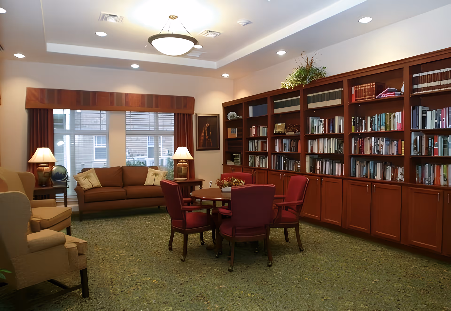 A well-lit common room with built-in bookshelves, a round table with red chairs, sofas, lamps, and large windows.