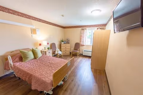 A private room in a skilled nursing facility featuring a hospital-style bed with a pink quilt and green pillows, two chairs, a small dresser, a window with curtains, a wall-mounted television, and wooden flooring.
