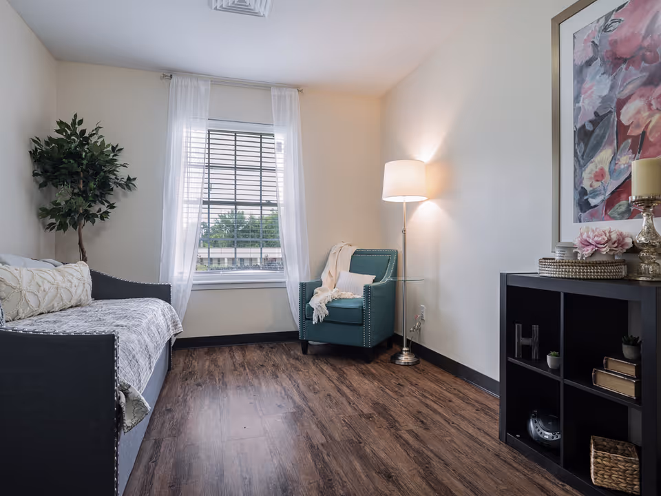 Bright furnished sitting room with a daybed, teal armchair, floor lamp, window with sheer curtains, potted plant, and shelving on wood floor.