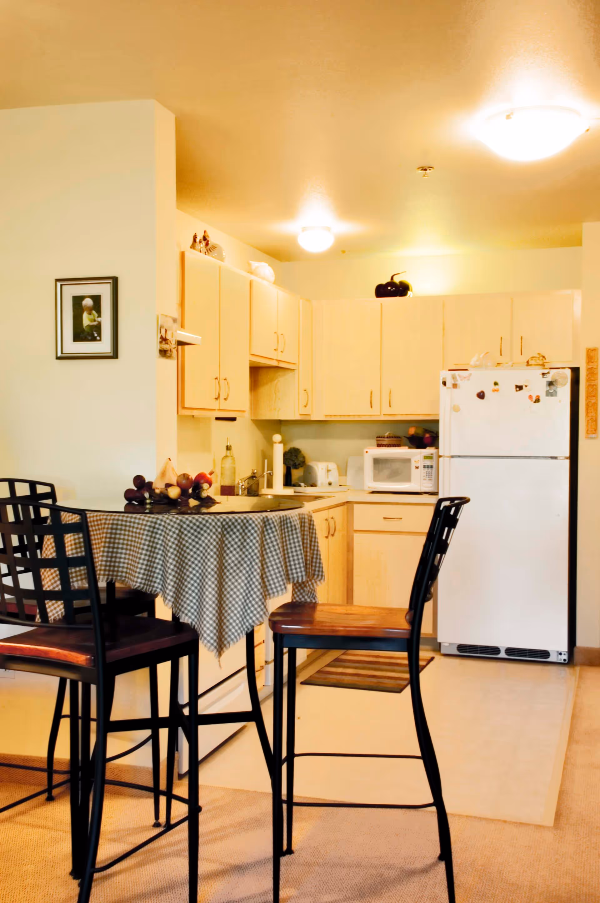Interior view of a kitchen area with light wood cabinets, a white refrigerator, microwave, and small appliances on the countertop. In the foreground, there is a round table covered with a checkered tablecloth and surrounded by three high chairs with black metal frames and wooden seats. The room is warmly lit with ceiling lights.