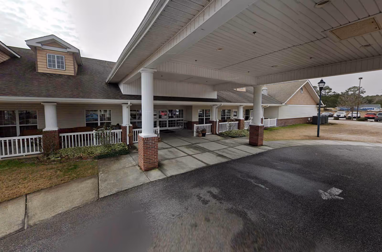 Exterior view of the entrance to Rockwell Park Rehabilitation and Healthcare Center showing a covered drop-off area supported by white columns with brick bases, a paved driveway, and a parking area with several cars in the background.