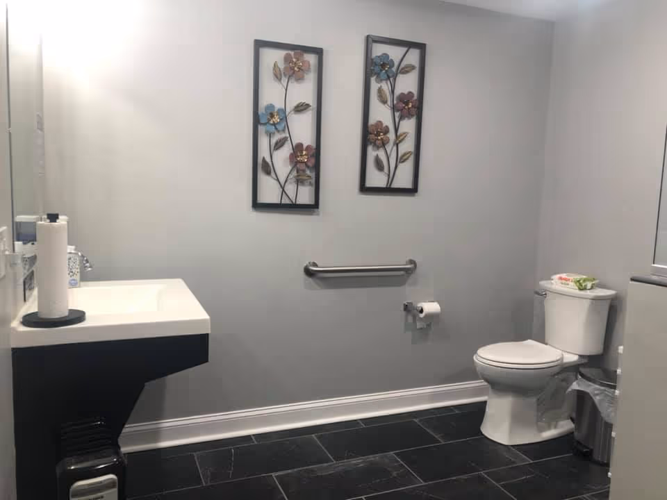 A clean accessible bathroom with a wall-mounted sink, toilet, grab bar, decorative floral wall art, and dark tiled floor.