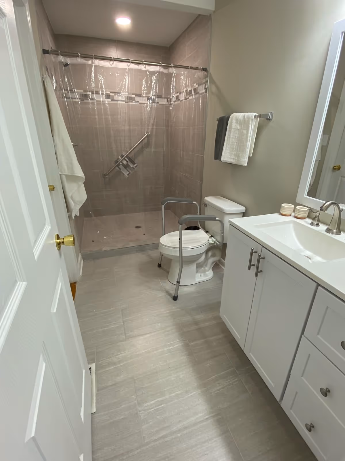 A clean, accessible bathroom with a walk-in shower featuring a grab bar and a clear shower curtain. There is a toilet with safety rails around it, a white vanity with a sink and faucet, a mirror above the sink, and towels hanging on a towel rack. The floor is tiled in a light gray color.