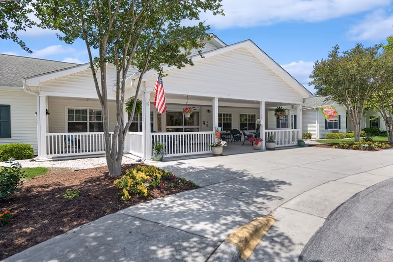 Exterior view of a white single-story building with a covered porch, white railings, and an American flag hanging near the entrance. There are potted plants and hanging flower baskets on the porch, with trees and landscaped flower beds surrounding the building under a blue sky with some clouds.