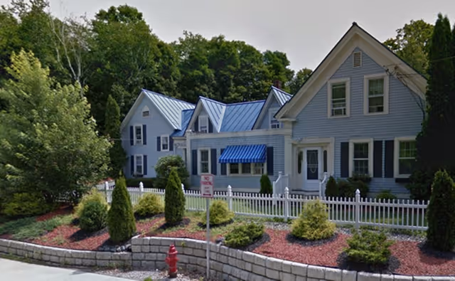 Exterior view of a light blue two-story house with white trim and a blue metal roof, surrounded by a white picket fence and landscaped with bushes and small trees. There is a red fire hydrant and a 'No Parking' sign near the curb in front of the house.