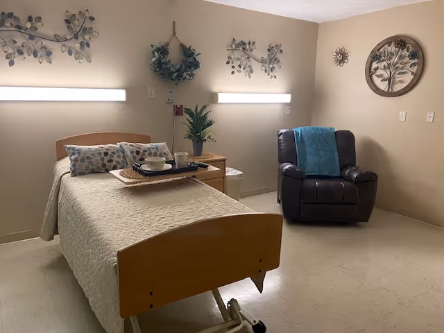 A simple, clean bedroom in a senior living facility with a single hospital-style bed covered with a beige quilt and a decorative pillow. A tray with cups and a bowl is placed on the bed. Next to the bed is a wooden nightstand with a small green plant. On the right side of the room, there is a dark brown recliner chair with a blue throw blanket draped over it. The walls are decorated with metal leaf and flower art pieces, and two horizontal lights are mounted above the bed.