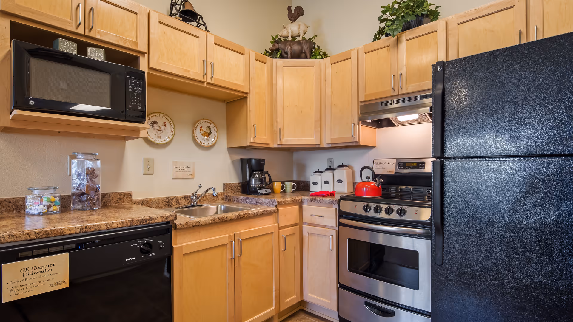 A compact kitchen with light wood cabinets, a black microwave mounted above the counter, a double sink, a black dishwasher, a stainless steel oven with a red kettle on top, and a black refrigerator. The countertop has jars with candy and a coffee maker. Decorative plates and small signs are on the wall, and there are animal figurines and a plant on top of the cabinets.