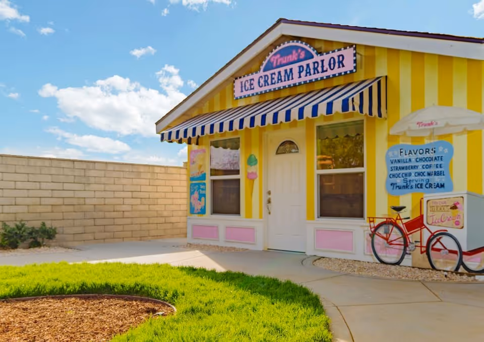 Yellow-and-pink small ice cream parlor storefront with a blue-and-white awning and a bicycle cart in a sunny courtyard.