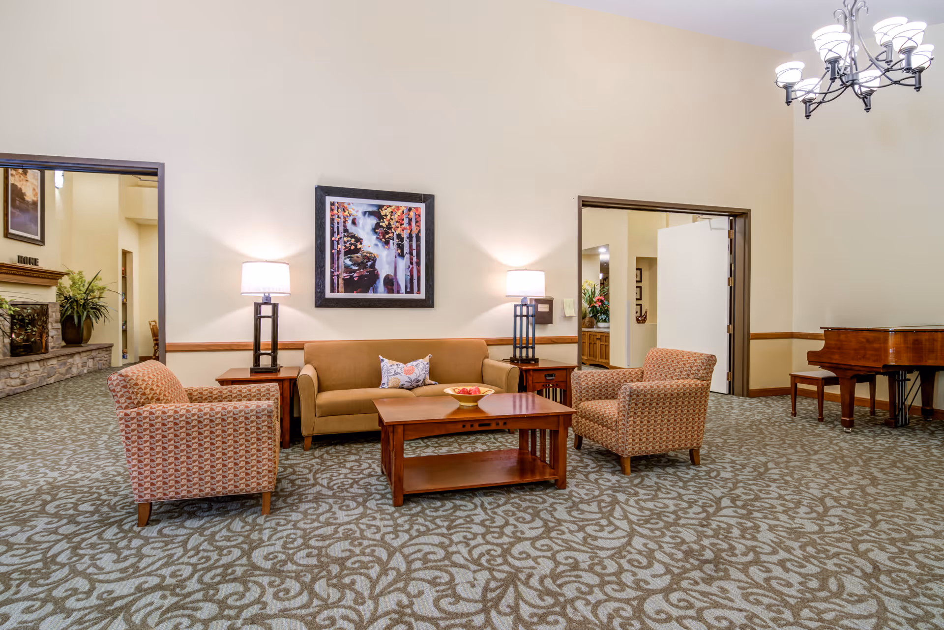 A senior living facility common area with a beige sofa, two patterned armchairs, a wooden coffee table with a bowl of apples, two table lamps on side tables, a framed painting of a waterfall on the wall, a grand piano in the corner, and a patterned carpet. The room has cream-colored walls and a chandelier hanging from the ceiling.