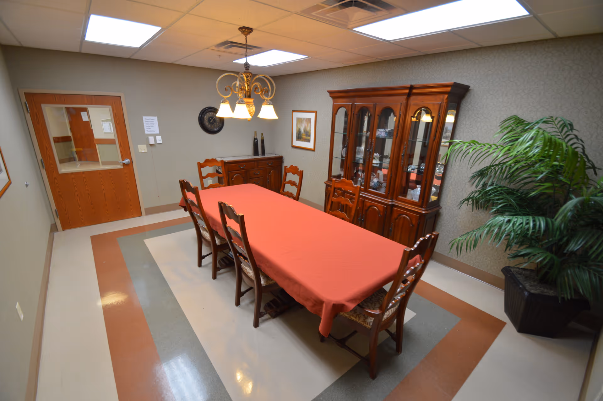 A dining room with a long rectangular table covered with a red tablecloth and six wooden chairs around it. There is a wooden china cabinet filled with dishes against the wall, a sideboard with decorative items, a framed picture, a wall clock, and a large potted plant in the corner. The room has a tiled floor with a patterned border and a ceiling light fixture with three lamps.