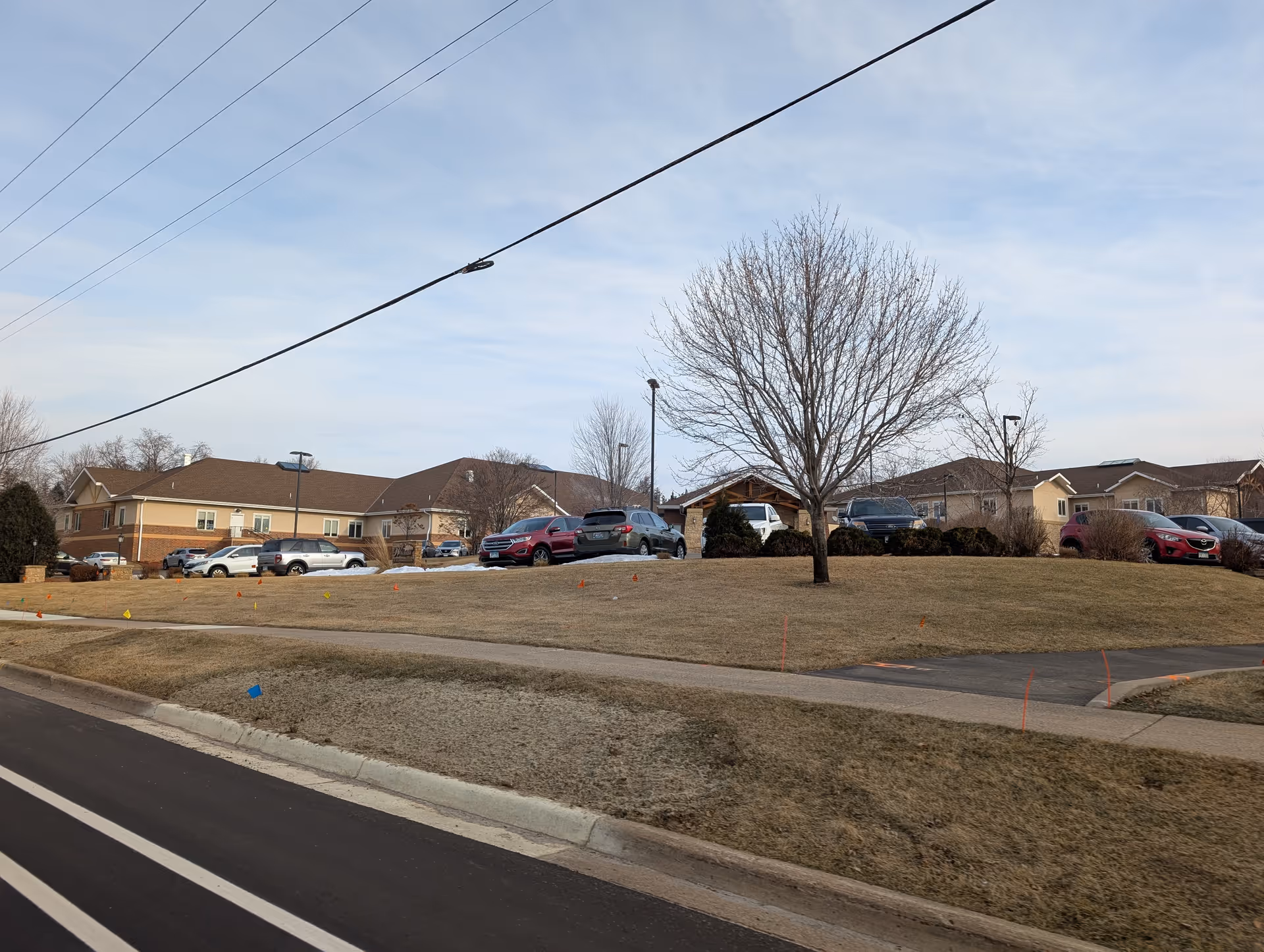 Exterior view of a senior living facility named Wealshire of Bloomington showing a large grassy area with a sidewalk, several parked cars, leafless trees, and a building with a brown roof under a partly cloudy sky.