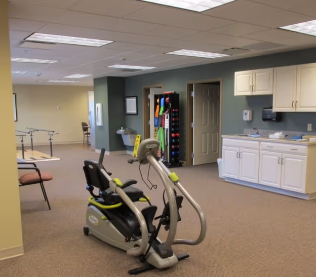 A spacious interior therapy room with a recumbent exercise machine, parallel walking bars, storage cabinets, and therapy equipment.