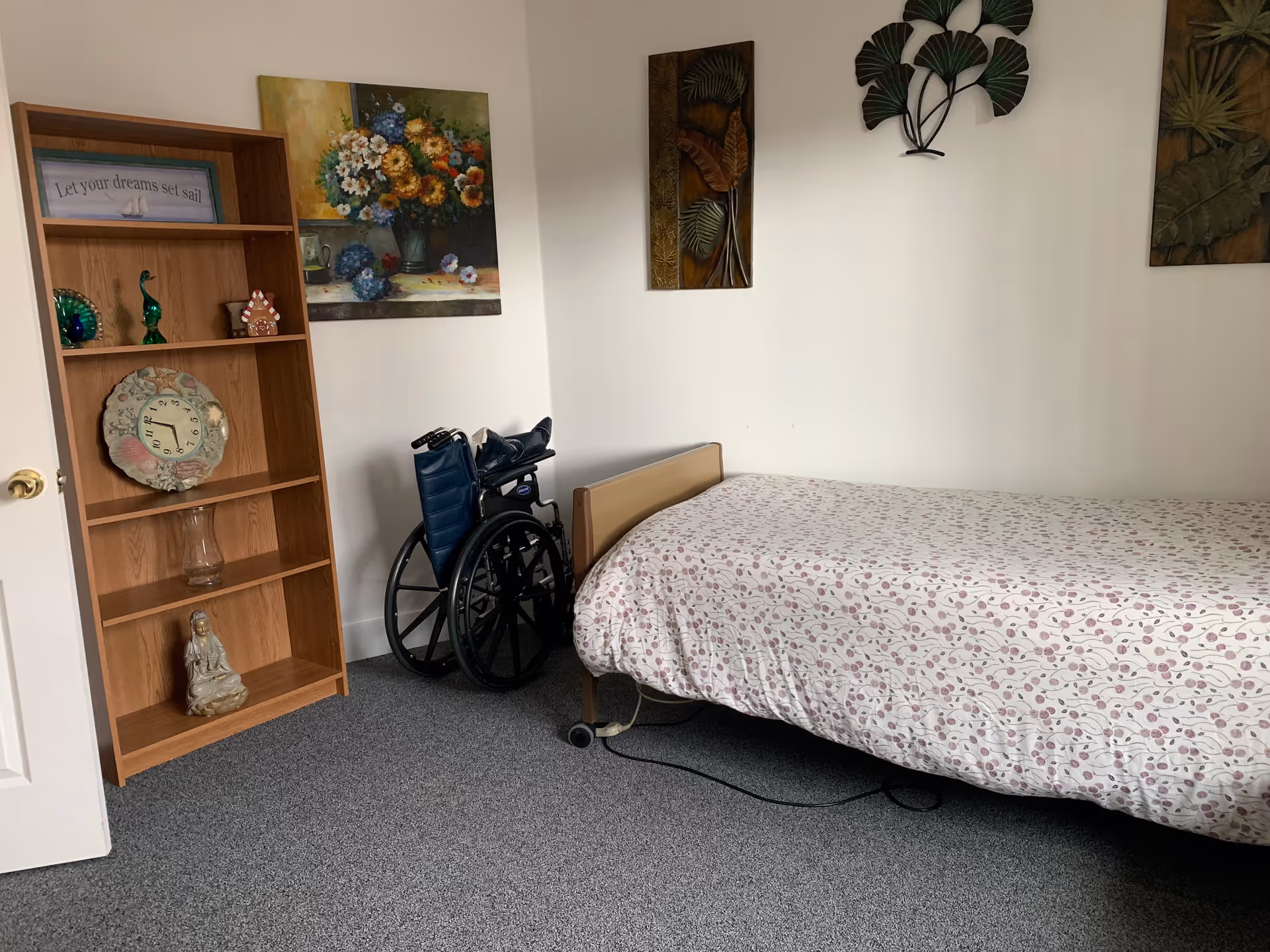 A simple bedroom with a single bed covered in a white bedspread with a pink floral pattern. Next to the bed is a blue wheelchair. A wooden bookshelf stands against the wall with decorative items including a clock, figurines, and a framed sign that reads 'Let your dreams set sail'. The walls are decorated with floral and leaf-themed artwork.