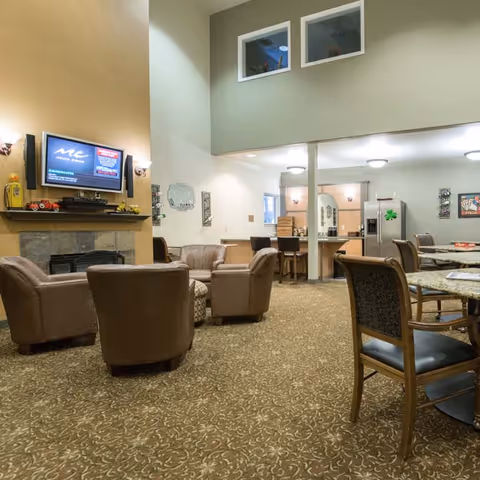 A spacious senior living common area with a high ceiling, featuring a seating arrangement of four brown leather chairs around a small round table near a fireplace with a mounted TV above it. In the background, there is a kitchen area with a refrigerator and bar stools, and a dining area with tables and chairs. The room has patterned carpet flooring and neutral-colored walls.