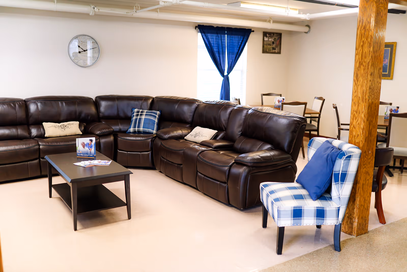 A cozy seating area in a senior living facility featuring a large dark brown leather sectional sofa with decorative pillows, a black coffee table with brochures, a blue and white checkered armchair with a blue cushion, and a wooden support column. In the background, there are dining tables with chairs and a window with blue curtains.