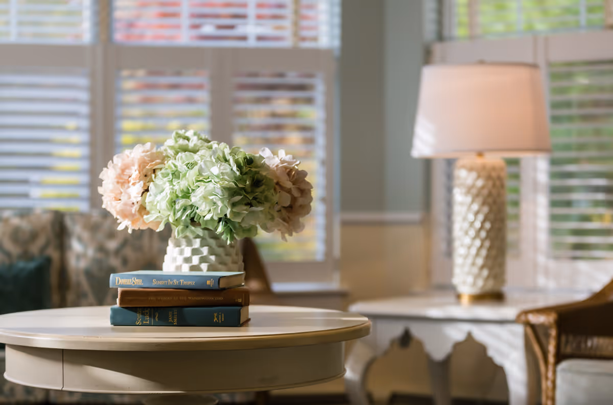 A round table in a sunlit living room holds a vase of hydrangeas atop stacked books with a lamp and window blinds in the background.