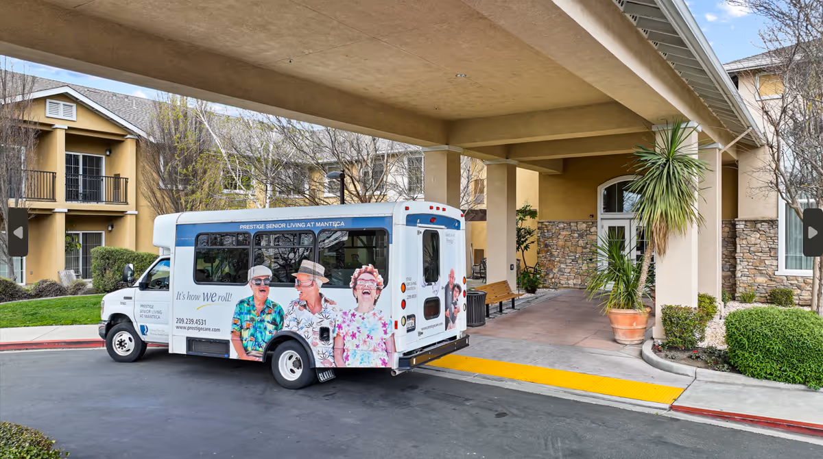 A small shuttle bus parked under a covered entrance of a senior living facility. The bus has colorful images of smiling elderly people and text that reads 'Prestige Senior Living at Manteca' and 'It's how WE roll!'. The building has stone and beige walls with some potted plants and bushes nearby.