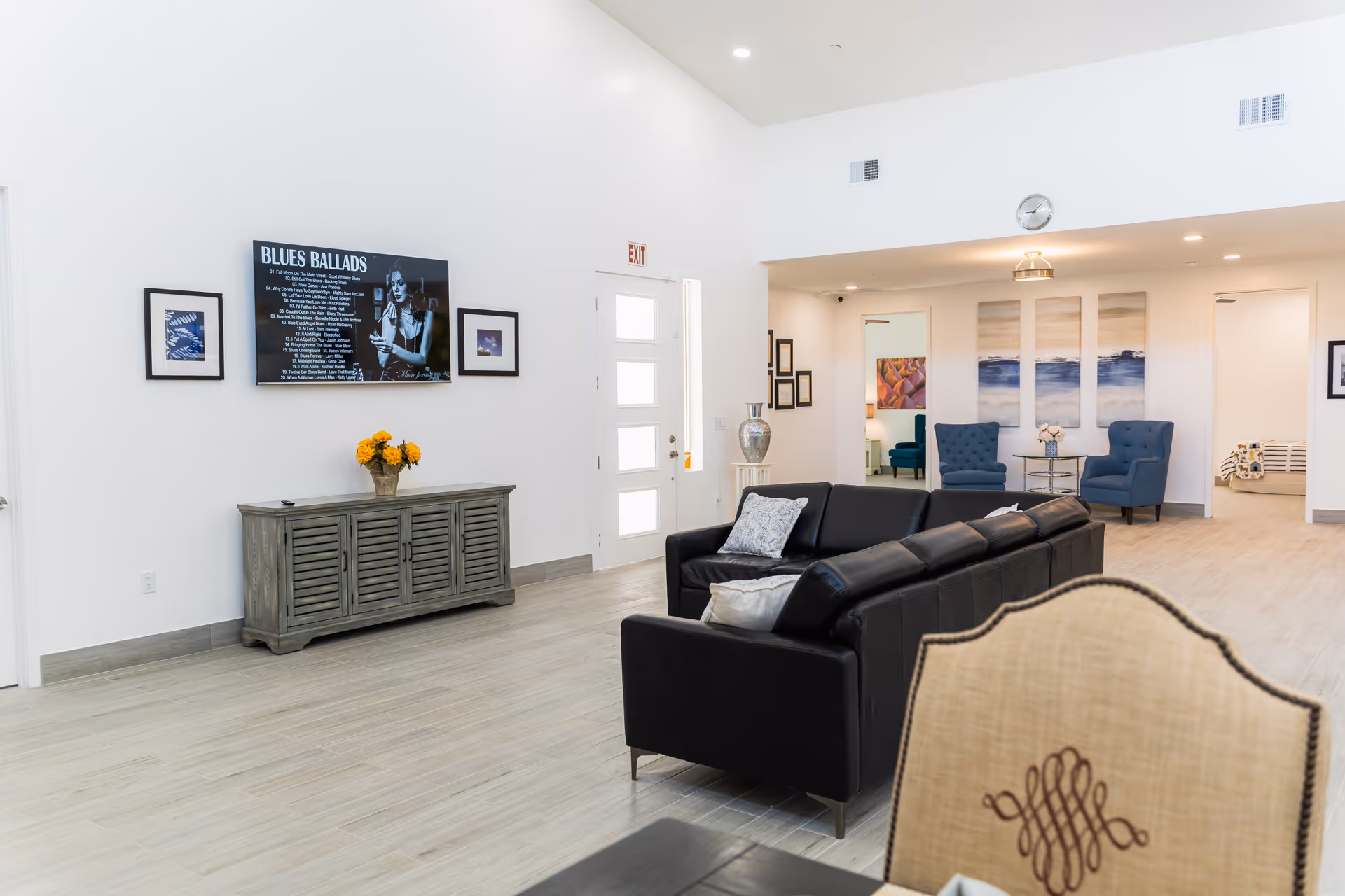 A bright and spacious living room area in Sunrise Care Home Shea featuring a black leather sectional sofa with pillows, two blue armchairs with a small round table between them, and a wooden sideboard with a vase of yellow flowers. A flat-screen TV mounted on the wall displays a 'Blues Ballads' playlist. The room has light wood flooring, white walls, and high ceilings with recessed lighting. Various framed artworks and a clock are visible on the walls.