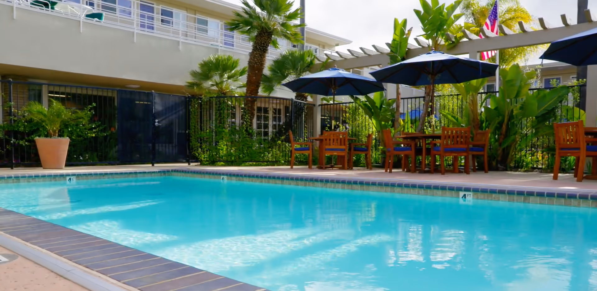 Outdoor swimming pool area with clear blue water, surrounded by a patio with wooden tables and chairs under blue umbrellas. There are palm trees and other greenery around the pool, with a building and a black fence in the background.