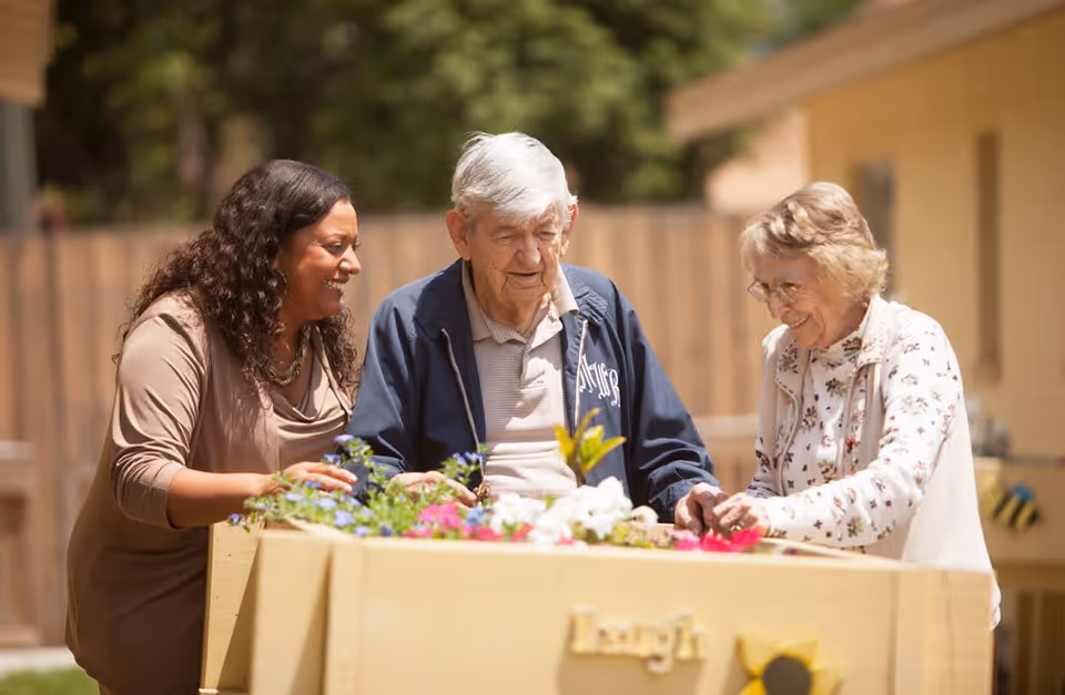 Two elderly individuals and a caregiver are enjoying gardening together outdoors, tending to a raised garden bed filled with colorful flowers. They are smiling and engaged in the activity, with a wooden fence and building in the background.
