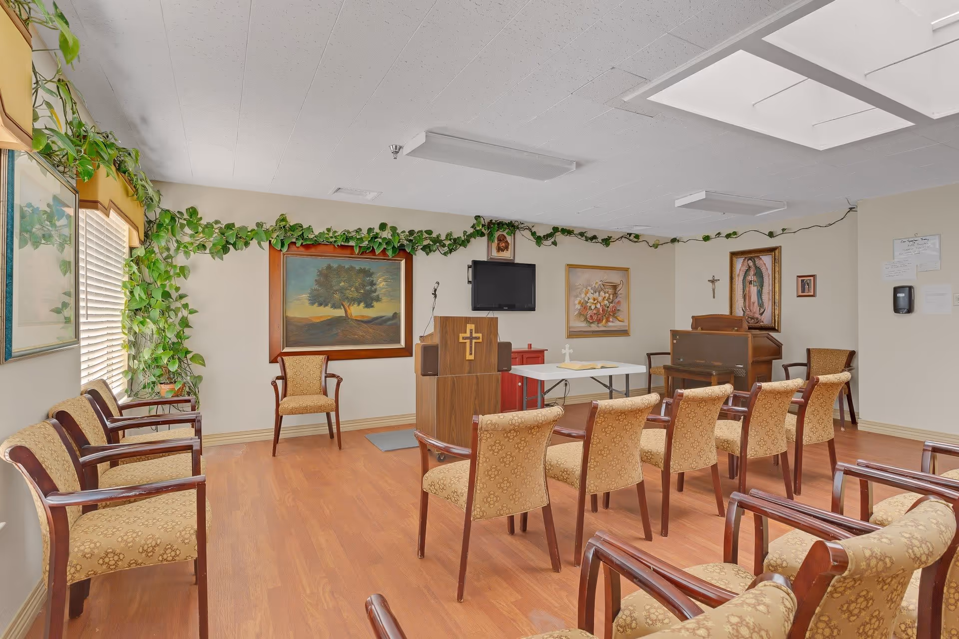 A small chapel or prayer room with wooden chairs arranged in rows facing a wooden podium with a cross. The room has light-colored walls decorated with paintings, a crucifix, and green leafy garlands along the top edges. There is a piano in the corner and a flat-screen TV mounted on the wall behind the podium. The floor is wood, and there is a window with blinds on the left side.