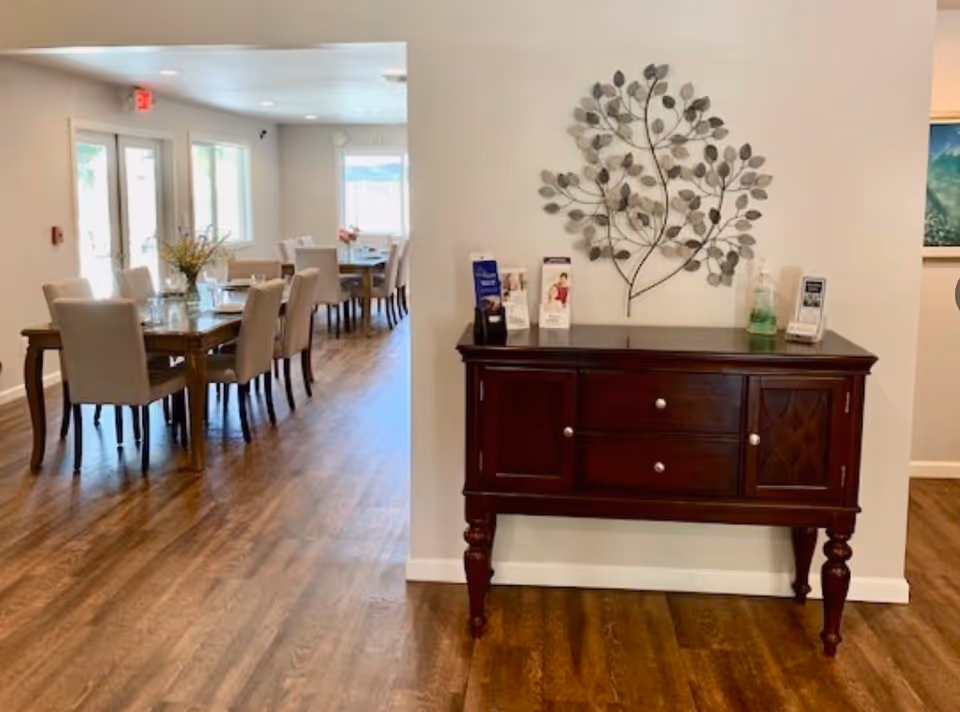 Interior view of a senior living facility dining area with wooden floors and beige upholstered chairs around wooden tables. A dark wooden sideboard with brochures and a decorative metal wall art piece shaped like branches with leaves is visible in the foreground.
