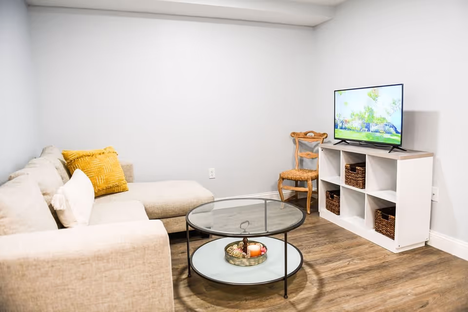 A cozy living room with a beige sectional sofa adorned with yellow and white pillows, a round glass coffee table with decorative items, a wooden chair with a woven seat, and a TV on a white cubed shelf unit against a light gray wall.