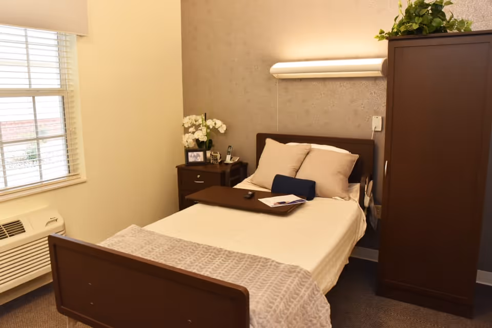 A neatly made single bed in a furnished bedroom with a nightstand, wardrobe, window, and overhead light.