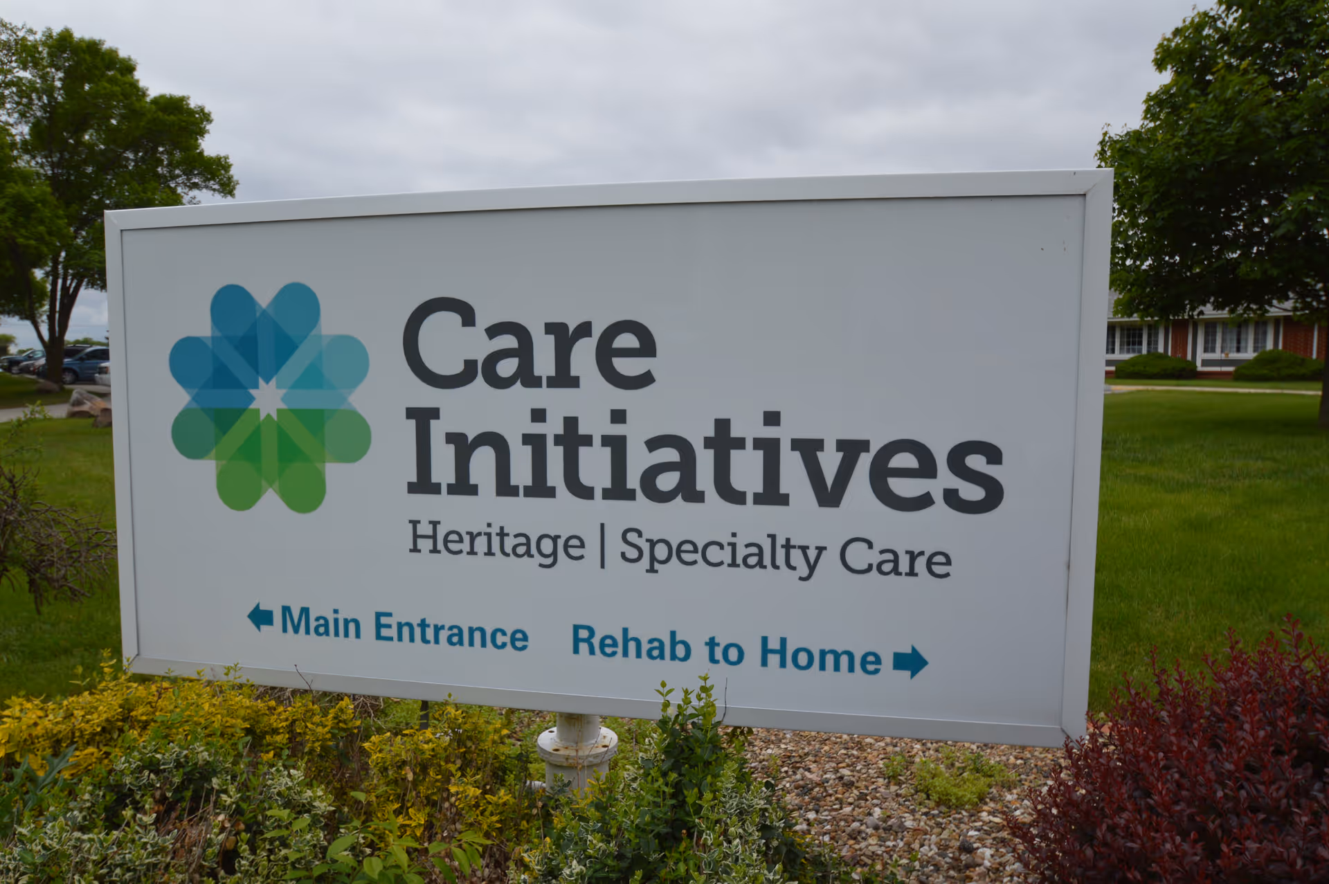 A freestanding outdoor sign reading 'Care Initiatives Heritage | Specialty Care' with arrows for Main Entrance and Rehab to Home in front of a grassy lawn.