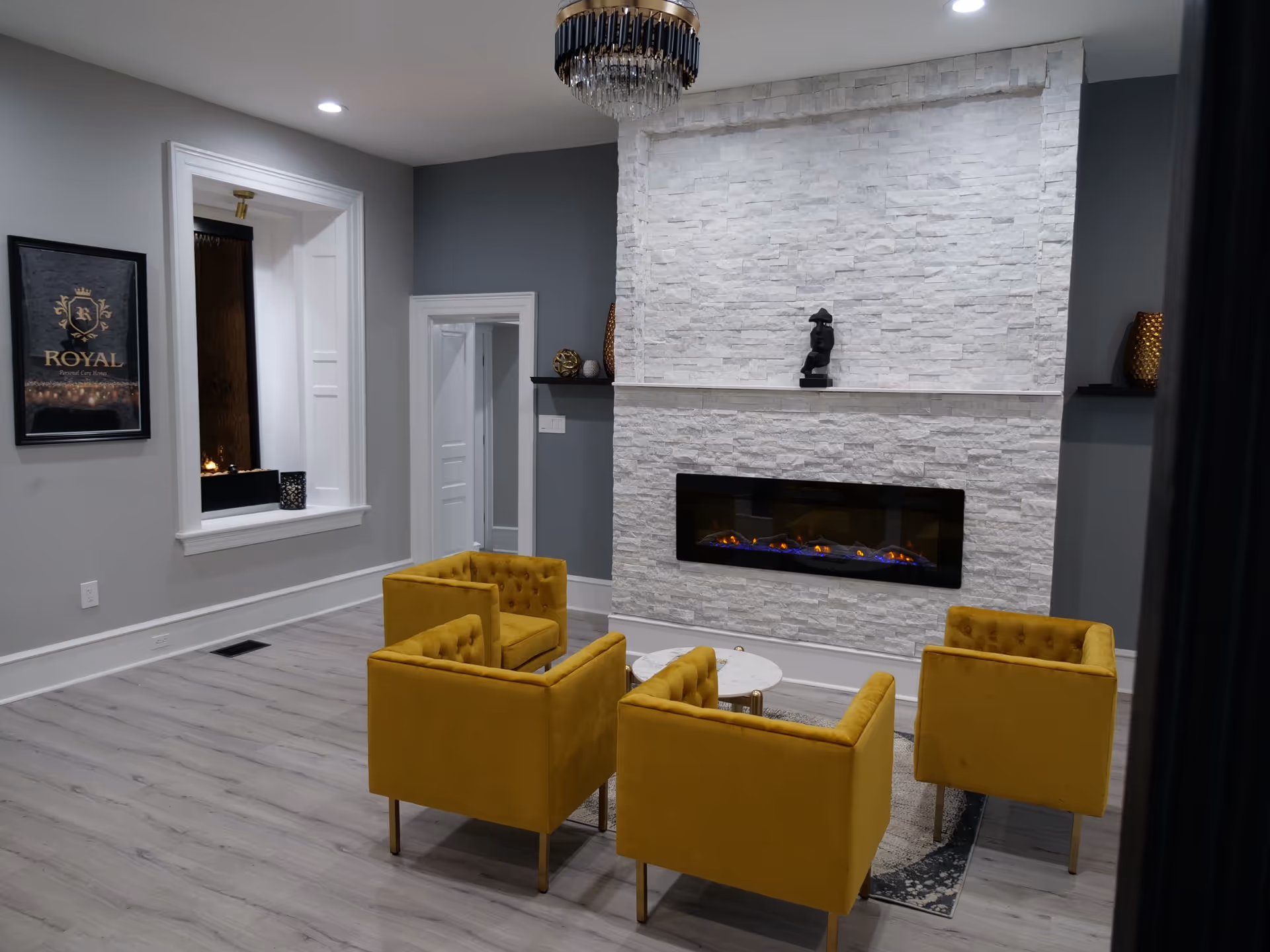 A modern living room with four mustard yellow armchairs arranged around a small round white table on a rug. The room features a white stone accent wall with a built-in electric fireplace and a black sculpture on a narrow shelf above it. The walls are painted gray, and there is a framed Royal Personal Care Homes logo on one wall. The floor is light wood, and a chandelier hangs from the ceiling.