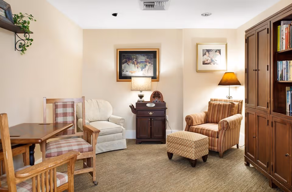 A cozy living room with beige walls and carpeted floor featuring a wooden table with two plaid cushioned chairs on the left, a cream-colored armchair in the center, a striped armchair with a matching ottoman on the right, a wooden cabinet with books, two framed pictures on the wall, and two table lamps providing warm lighting.