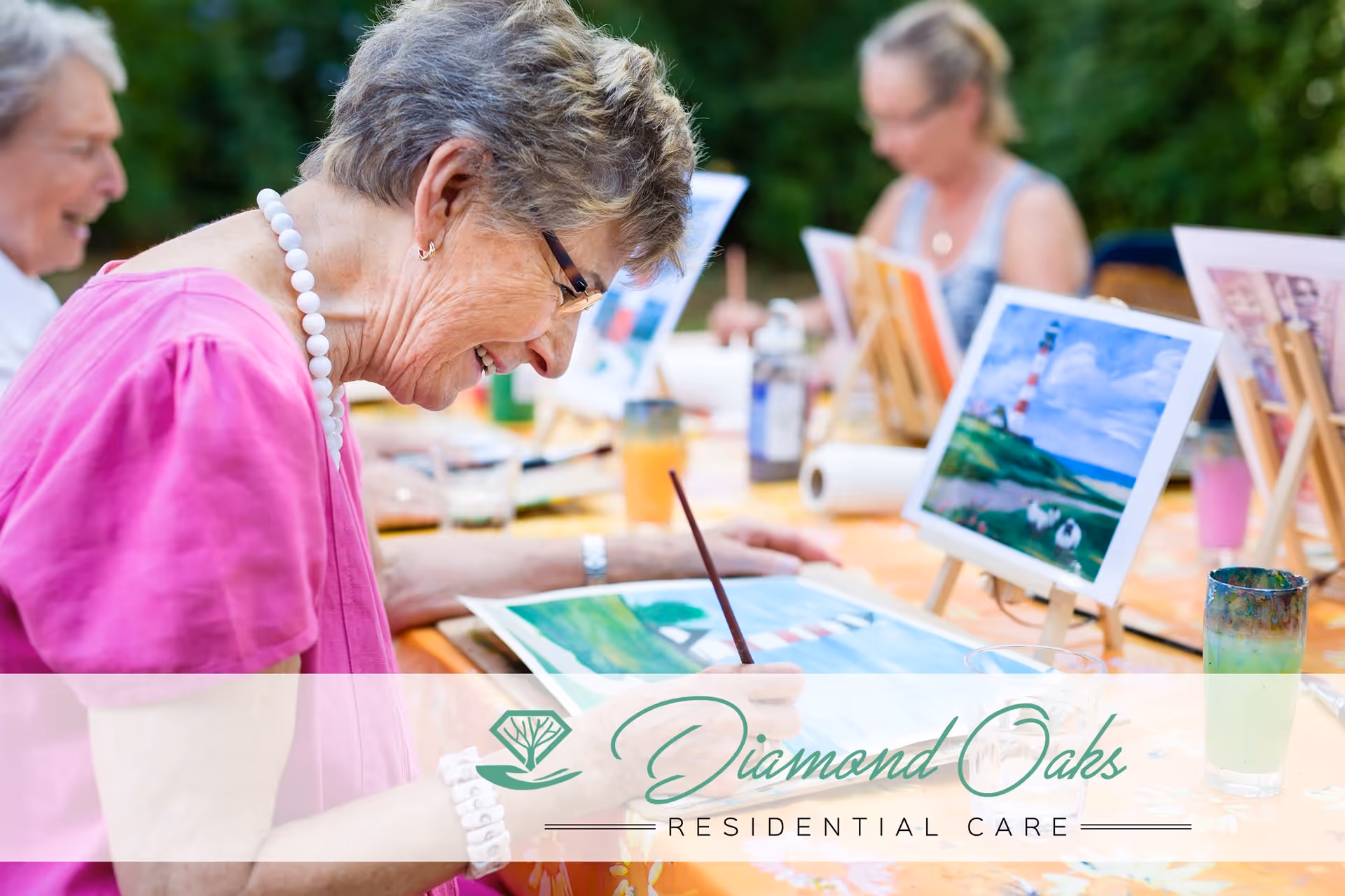 Elderly women engaged in a painting activity outdoors, sitting at a table with easels and colorful paintings, surrounded by art supplies and greenery in the background.