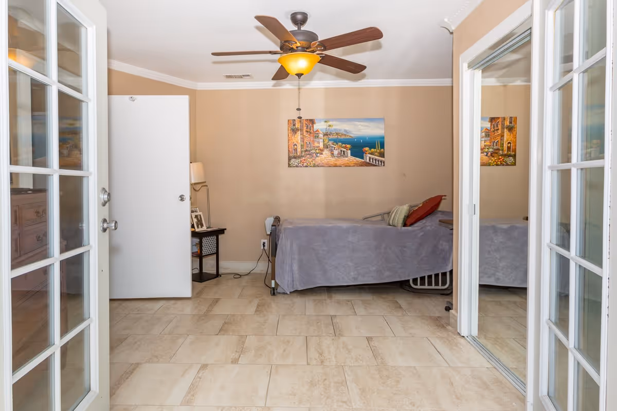 A small bedroom with beige walls and tiled floor, featuring a single bed covered with a gray blanket and pillows. There is a ceiling fan with a light fixture above, a side table with a lamp and framed photos, a white door slightly open, and a large mirror on the right wall reflecting part of the room. A colorful painting of a seaside village hangs on the wall above the bed.